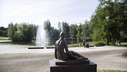 Sorsapuiston istuvan naisen patsas takaapäin kuvattuna. Patsas katsoo kohti suihkulähdettä.