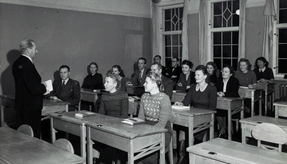 Luokkahuone, jossa opiskelee ihmisiä pulpeteissa. Etualalla on opettaja.