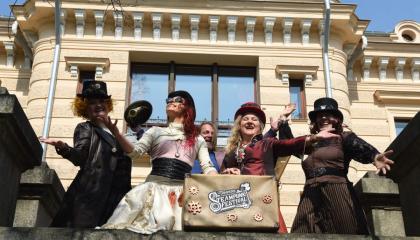 Viisi esiintyjää rooliasuissa vanhan rakennuksen edessä. Kaksi esiintyjistä pitelee vanhaa matkalaukkua, jossa lukee Tampere Steampunk Festival