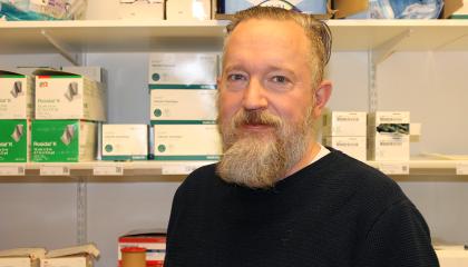 Parrakas mies hymyilee vienosti varastohyllyjen edessä. 