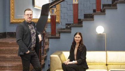 Heikki Aittala seisoo portaiden edessä ja Peppiina Inervo istuu sohvalla Tuulensuun palatsin aulassa, molemmat katsovat kameraa kohti hymyillen. 