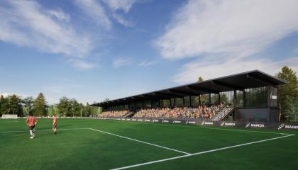 Jalkapallokentällä kaksi pelaajaa ja taustalla kasomossa näkyy katsojia istumassa katoksen alla.