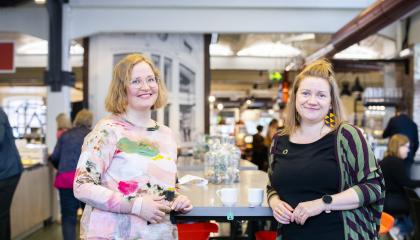 Kaksi henkilöä seisoo nojaten pöytään ja katsoo kameraan.