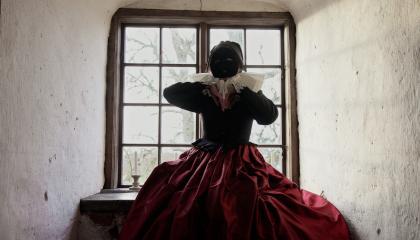 Myllynkivikaulukseen, mustaan paitaan ja punaiseen hameeseen pukeutunut hahmo istuu vanhan linnan ikkunan edessä. Hahmolla on musta naamio kasvoilla.