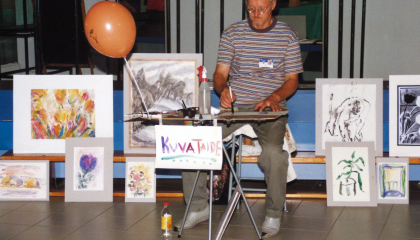 Henkilö istuu pöydän ääressä piirtämässä. Hänen ympärillään on erilaisia tauluja.