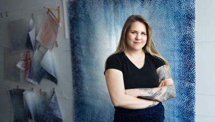 Mustahameinen, pitkähiuksinen nainen seisoo sinisävyisen seinälle ripustetun taideteoksen edessä. 