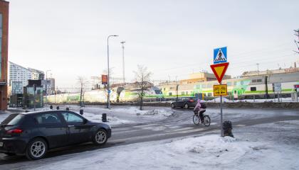 Pakkahuoneenaukion ja Ratapihankadun liittymä pohjoisesta kuvattuna, auto ja pyöräilijä ylittää tietä, juna taustalla.