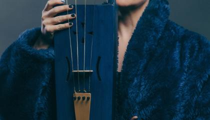 Lyhythiuksinen nainen pitelee siniseksi maalattua jouhikkoa puoliksi kasvojensa edessä. Naisella on sininen turkistakki.