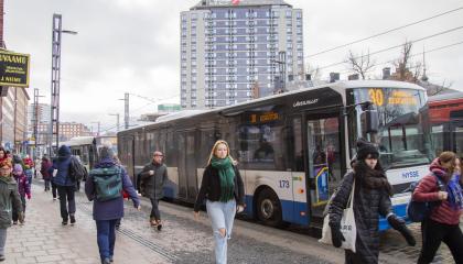 Useita ihmisiä kadulla kävelemässä ja taustalla bussi sekä korkea talo.