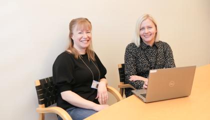 Kaksi henkilöä istuu pöydän ääressä tietokoneen kanssa ja katsovat hymyillen kameraan.