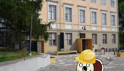 Taustalla vaalea vanha kivirakennus, jonka edessä mukulakivipiha. Edustalla piirretty Rulla-hahmo seisoo päässään keltainen rakennuskypärä.