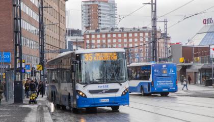 Tampereen keskustassa kaksi sinivalkoista kaupunkiliikenteen bussia toinen menossa ja toinen tulossa. Kadulla ihmisiä bussipysäkillä ja taustalla kaupunkinäkymää.
