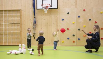 Kolme lasta ja päiväkodin työntekijä heittelevät palloja päiväkodin liikuntasalissa.