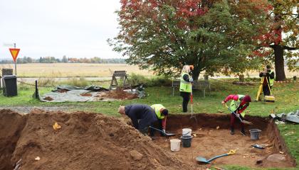Ihmisiä arkeologisilla kaivauksia. Maahan on kaivettu kuoppa, jossa kolme henkilöä lapioi hiekkaa ämpäreihin. Maisema on syksyinen.