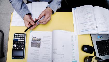 Ylhäältä kuvattu opiskelijan työpöytä, jossa näkyy kirja, laskin ja kynä opiskelijan kädessä.