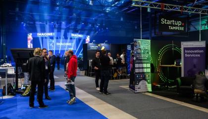 Messuhallissa tapahtumaständejä ja ihmisiä, taustalla päälava ja valonheittimiä. 