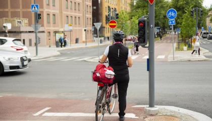 Henkilö seisoo selin liikennevaloissa taluttaen polkupyörää.