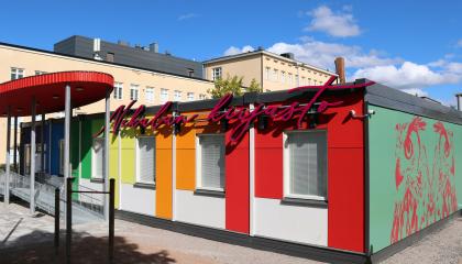 Nekalan konteista tehty värikäs kirjasto ja sen edustalla oleva ovaalinmuotoinen katos. Taustalla näkyy Nekalan koulurakennusta. 