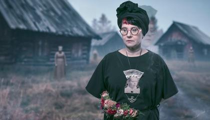 Naisella on päällään tummat vaatteeet. Kaulassa roikkuu suuri koru, jossa on mustavalkoinen kasvokuva.