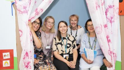 Viisi naista istumassa valkoisten, vaaleanpunakuvioisten verhojen välissä.