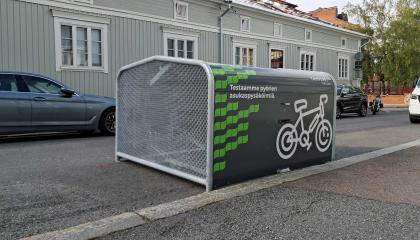 Pyörien  säilytyshangaari kadun reunassa, toisella puolella puutalo.