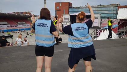 Kaksi henkilöä seisoo Ratinassa siniset liivit päällään. Liiveissä lukee lukee &quot;miten menee&quot;.