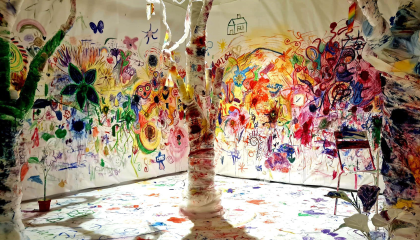 Etualalla kolme valkoisella hallaharsolla verhoiltua käsin tehtyä puuta. Hallaharsoihin on maalattu eri väreillä. Puiden taustalla valkoisella paperoitu seinä, johon myös maalattu eri väreillä. 