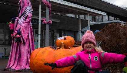 OIkealla leikkivä lapsi, vasemmalla taaempana pinkkeihin kaapuihin pukeutunut hahmo. Välissä puhallettava iso kurpitsa.