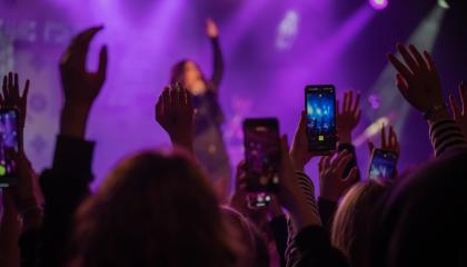 Yleisö kädet ilmassa. Etualalla puhelin, joka kuvaa lavaa, jossa näkyy esiintyjä.