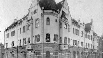 Mustavalkoisessa valokuvassa iso nelikerroksinen rakennus, jonka alatasossa on liiketilaa.