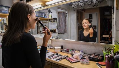 Nuori meikkaa itseään paksulla siveltimellä ison peilin edessä. Peilin alla olevalla pöydällä on erilaisia meikkejä.