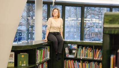 Nainen istuu ikkunan edessä kirjaston matalien kirjahyllyjen päällä ja hymyilee. 