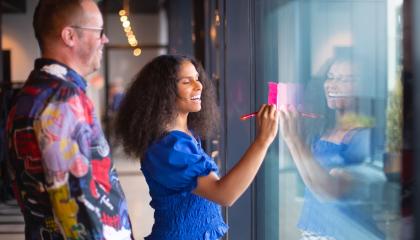 Kaksi henkilöä sivuttain toimiston aulassa, jossa isot ikkunat. Etumainnen kirjoittaa ikkunassa kiinni olevaan muistilappuun ja hymyilee. Taaempana oleva henkilö katselee ja hymyilee hänkin. 