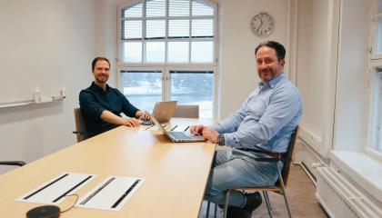 Kaksi miestä istuvat hymyillen kokouspöydän edessä läppärit kädessä. 