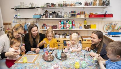 Kolme aikuista ja neljä pikkulasta ison pöydän ääressä. Lapset tutkivat keskittyneen näköisenä värikkäitä maalipurkkeja. Taustalla näkyy hyllykkö, jossa on siistissä järjestyksessä erilaisia kuvataidetarvikkeita.