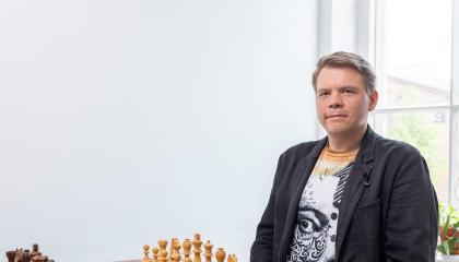 Mies istuu shakkilaudan äärellä, katsoo kameraan.