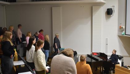 Henkilöt seisovat penkkiriveillä ja venyttelevät. Flyygelin vieressä istuu kuoronjohtaja.