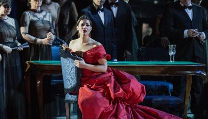 Nainen punaisessa iltapuvussa istuu tuolilla ja katsoo eteenpäin. Taustalla ihmisiä juhla-asuissa.