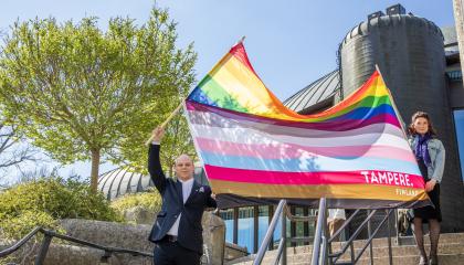 Kaksi henkilöä heiluttaa suurikokoista Tampereen omaa 13-raitaista pride-lippua pääkirjasto Metson rappusilla. 