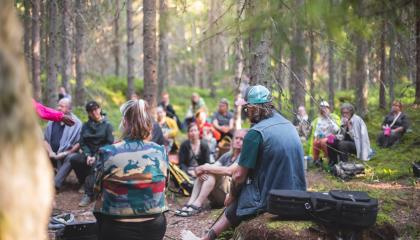 Iso joukko ihmisiä istuu Kintulammin metsässä kuuntelemassa musiikkiesitystä. 
