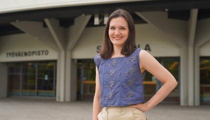 Henkilö seisoo rakennuksen edessä. Hän pitää kättään vyötäröllään.