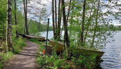 Järven rannalla soutuveneitä ja kapea hiekkapolku läpi lehtipuumetsän.