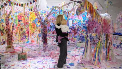 Nainen seisoo lapsi sylissään värikkäässä huoneessa, jossa valkoiselle taustalle on maalattu eri värein. Nainen ja lapsi katsovat suurta paperista tehtyä yksisarvista.