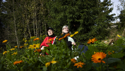 Kaksi henkilöä istuu keskellä yhteisöviljelmää, ympärillä kukkia, taustalla metsää.