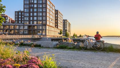 Kuvassa kerrostaloja, taivasta, kasveja edustalla ja järvmaisemaa taustalla.