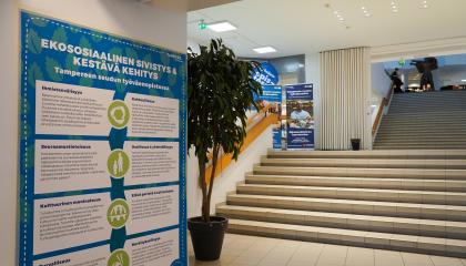 Seinällä juliste, jossa on paljon tekstiä. Taustalla huonekasvi ja portaikko.