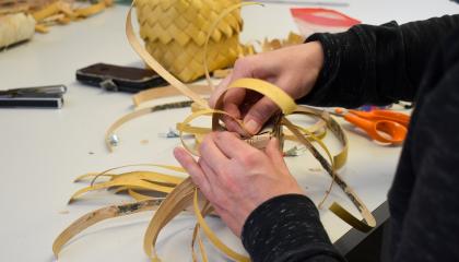 Henkilö sitoo tuohisuikaleita pöydän ääressä. Taustalla tuohirasia.