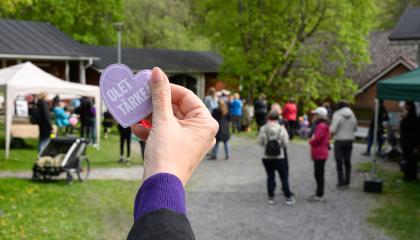 Kesäinen tapahtuma, jossa näkyy ihmisiä taustalla. Etualalla käsi, joka pitelee tarraa tekstillä "Olet tärkeä".