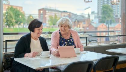 Kaksi henkilöä istuu kahvilassa ja katsoo hymyillen tablet-laitetta.