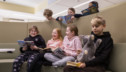 Koulun oppilaita istuu kirjojen ja pehmolelun kanssa ja nauraa. Osa oppilaista kurkkii ylhäältä sermien takaa.